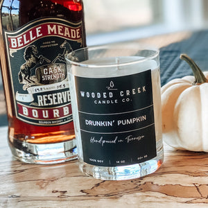 Hippies & Cowboys candle displayed with a bourbon bottle, pumpkin and a plant on a wooden table