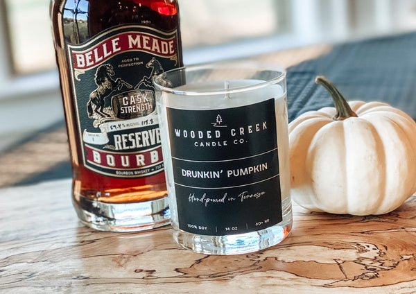 Hippies & Cowboys candle displayed with a bourbon bottle, pumpkin and a plant on a wooden table